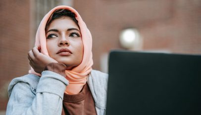 muslim female student daydreaming while sitting with laptop near city building