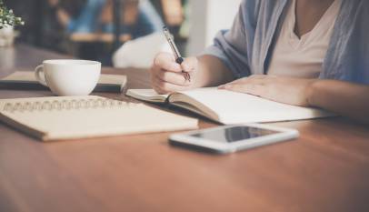 woman writing on a notebook beside teacup and tablet computer